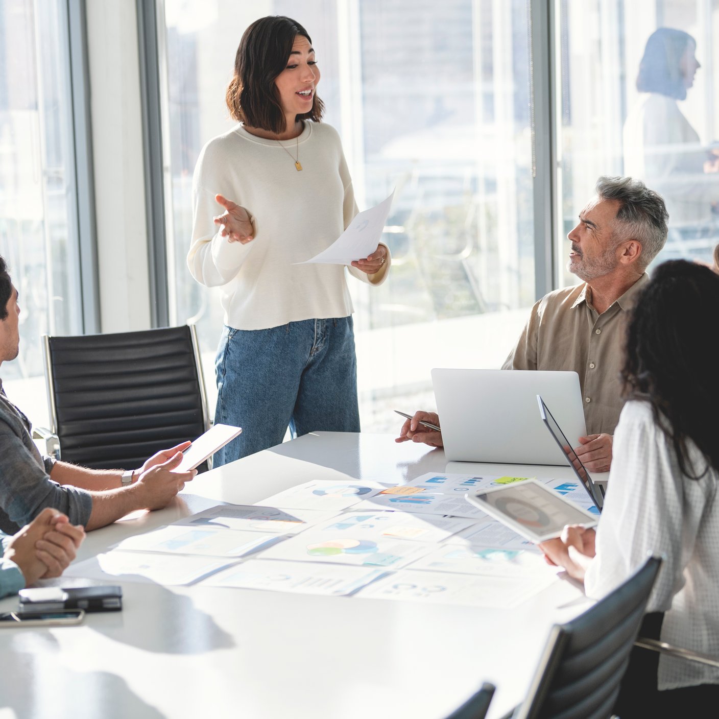 Confident woman giving a presentation with a group of people. She is holding a document. There is paperwork on the table with charts and graphs