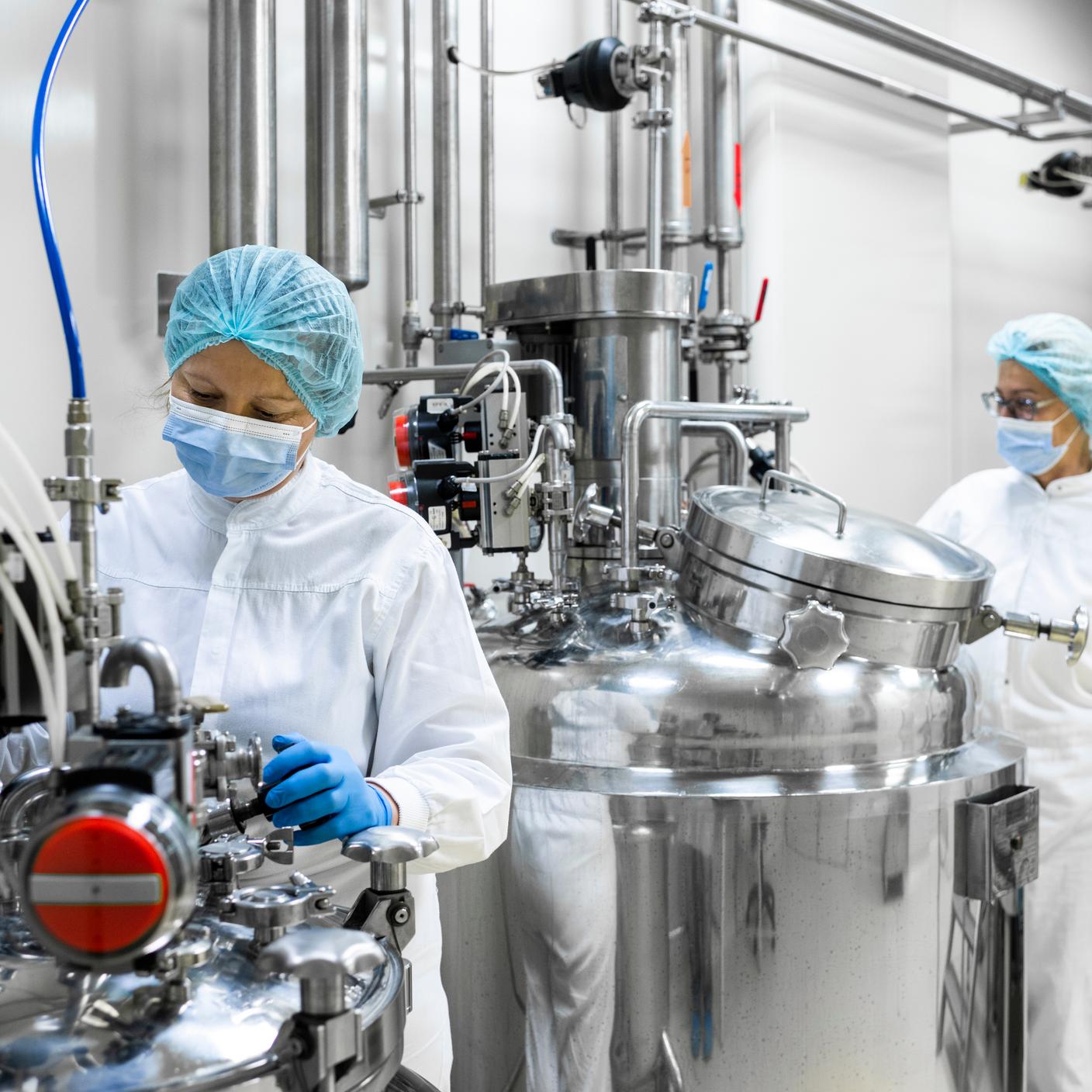 Two female employees in a laboratory wearing protective gloves, mask, cap and white suit seen standing each by one machine that is the part of the medicaments production during the working hours in a pharmaceutical manufacturing.