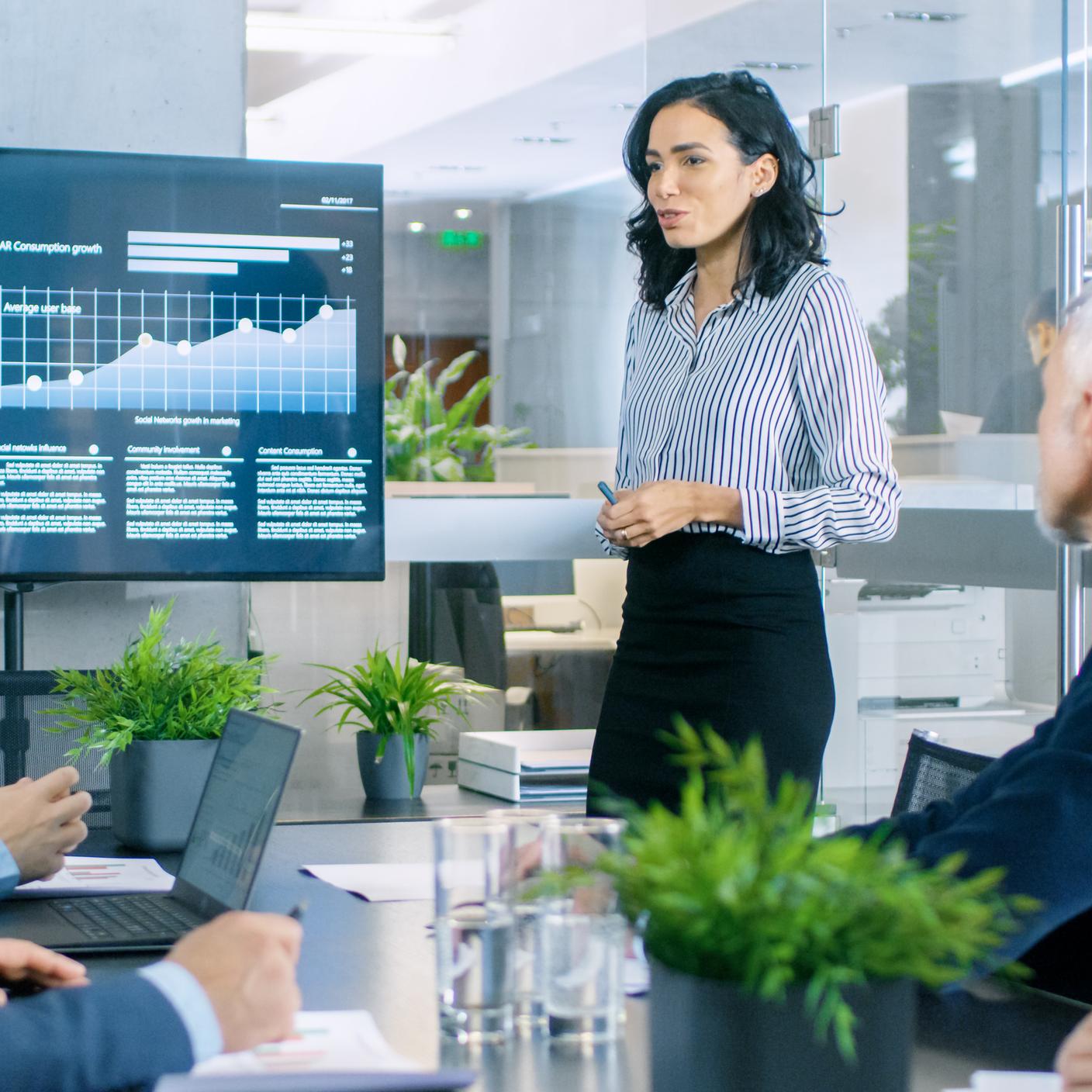 Businesswoman Gives Report/ Presentation to Her Business Colleagues in the Conference Room, She Shows Graphics, Pie Charts and Company's Growth on the Wall TV.