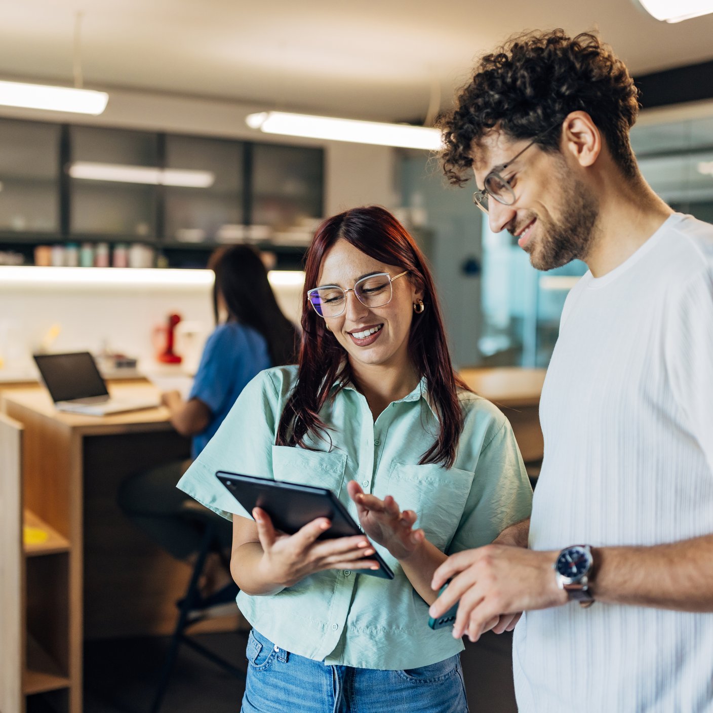 Two smiling business colleagues are looking at a digital tablet screen while standing in a hybrid workspace.