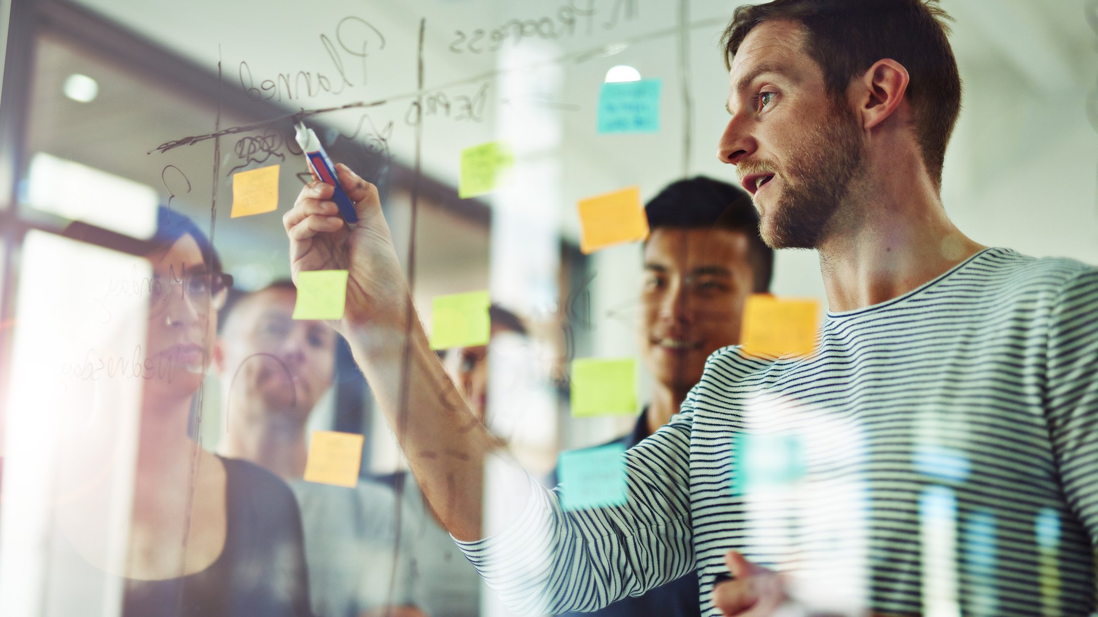 A person writes on a glass board with sticky notes while colleagues observe.